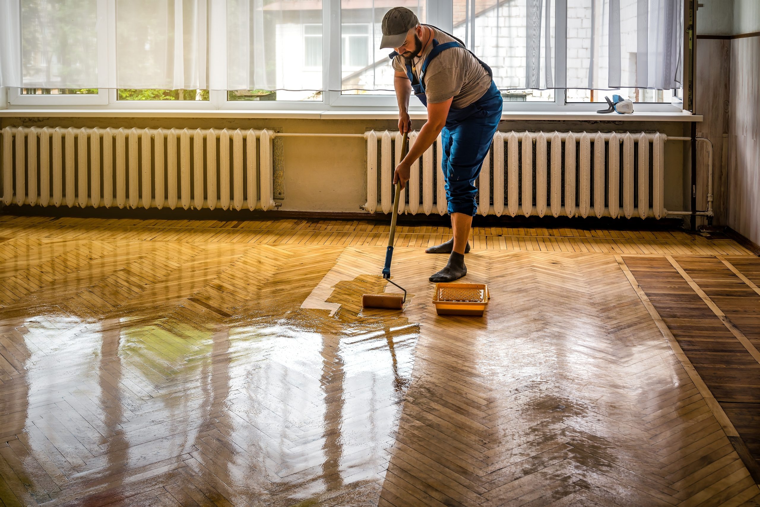 Parquet Floor Sanding in Egham with a technician applying lacquer to a freshly sanded oak herringbone parquet floor in a bright room.