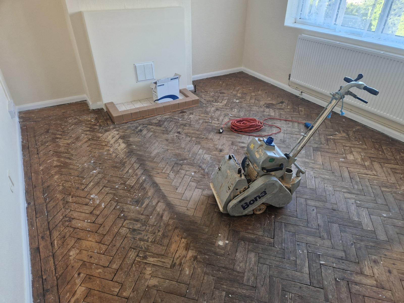 Parquet Floor Sanding in Egham underway on a worn oak herringbone parquet floor in a living room, with a professional sanding machine preparing the surface for restoration.