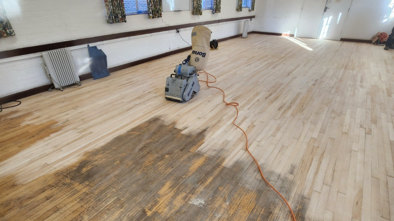Commercial Floor Sanding in Egham on a maple strip hardwood hall floor, showing a sanding machine in action and the contrast between old and newly sanded timber.